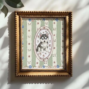 May include: A framed print featuring a watercolor illustration of a Siberian Husky puppy. The puppy is in an oval frame, set against a green and white striped background with floral accents. The frame is gold-colored.