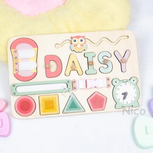 May include: Wooden educational toy with the name "DAISY" spelled out in colorful letters. Features a clock, shapes, a zipper, a buckle, and an owl. The toy is designed for early learning and sensory exploration.