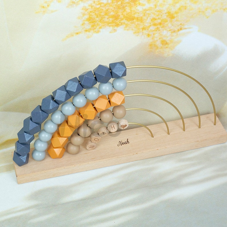 K&ouml;nnte beinhalten: Holz Regenbogen Spielzeug mit Perlen in Blaut&ouml;nen, Grau, Orange und Braun. Die Perlen sind auf einem goldenen Drahtbogen aufgef&auml;delt. Die Basis des Spielzeugs ist aus Holz und hat den Namen "Noah" eingraviert.