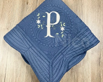 Puede incluir: Una manta acolchada azul con la letra "P" blanca bordada, rodeada de detalles florales. La manta presenta un diseño detallado y está doblada sobre una superficie de madera.