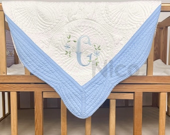 Könnte beinhalten: Eine weiße und hellblaue Babydecke mit einem gestickten Monogramm "G" in der Mitte. Die Decke ist mit einem kreisförmigen Muster gesteppt und hat einen hellblauen Rand.