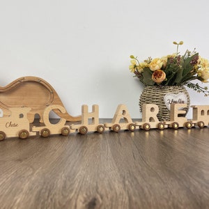May include: Wooden toy train with the name "CHASE" spelled out in individual train cars. The train is pulled by a wooden dinosaur money box.