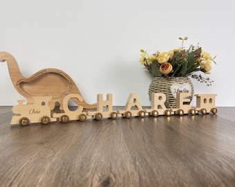 May include: Wooden toy train with the name "CHASE" spelled out in individual train cars. The train is pulled by a wooden dinosaur piggy bank.