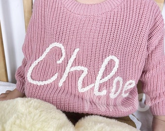May include: A pink knitted sweater with a white embroidered name "Chloe" on the front.