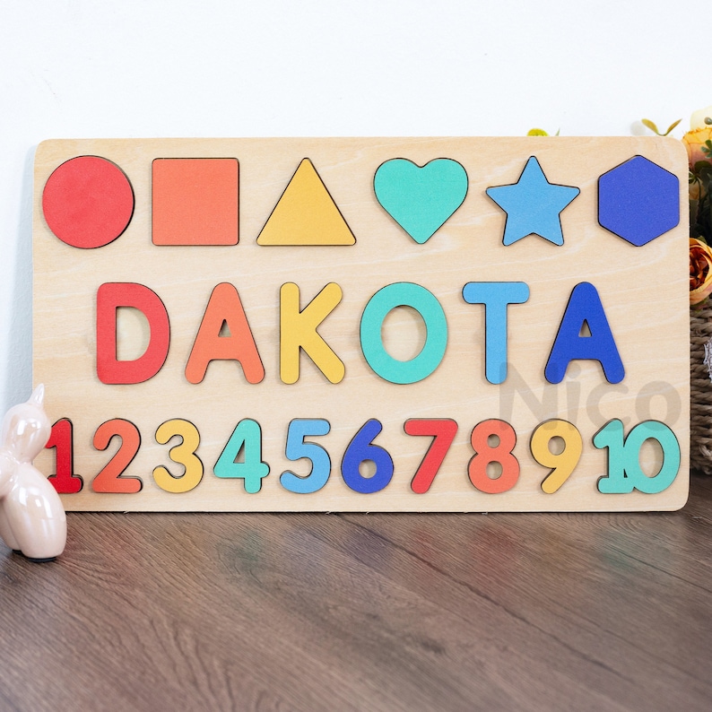 May include: Wooden puzzle with colorful shapes, letters, and numbers. The puzzle features the name DAKOTA, the numbers 1 through 10, and shapes including a circle, square, triangle, heart, star, and hexagon.