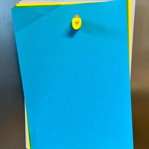 Puede incluir: Una pila de hojas de papel rectangulares de varios colores, incluyendo azul brillante, amarillo, rosa y gris claro. Un peque&ntilde;o im&aacute;n amarillo con forma de pato est&aacute; sujeto en la parte superior central de la hoja azul, manteniendo la pila unida.