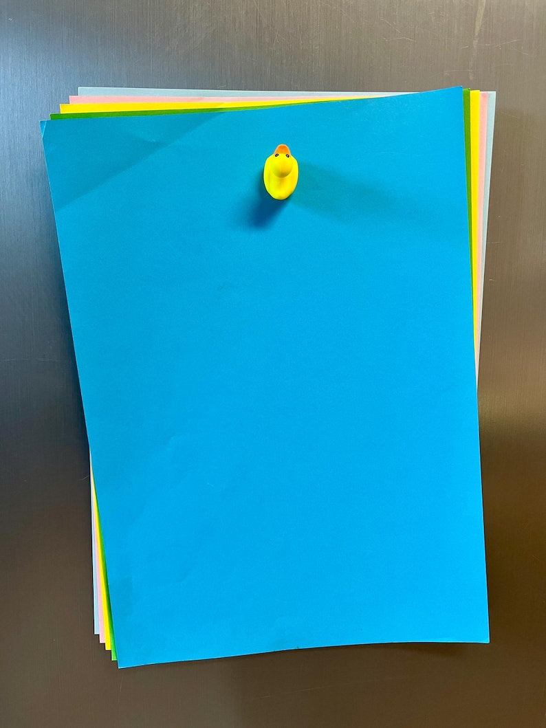 Puede incluir: Una pila de hojas de papel rectangulares de varios colores, incluyendo azul brillante, amarillo, rosa y gris claro. Un peque&ntilde;o im&aacute;n amarillo con forma de pato est&aacute; sujeto en la parte superior central de la hoja azul, manteniendo la pila unida.