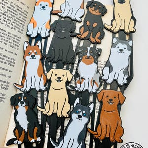 Può includere: Una collezione di segnalibri a tema cani. Ogni segnalibro presenta una razza di cane diversa in un design colorato e simile a un cartone animato. I segnalibri sono attaccati a nastri neri e sono ambientati sullo sfondo di un libro aperto. Il testo "MADE IN FRANCE" è visibile.