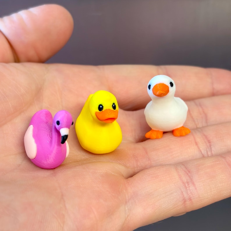 Puede incluir: Tres figuras en miniatura que incluyen un flamenco rosa brillante con alas de color rosa claro, un pato de goma amarillo vibrante con pico naranja y un patito blanco con pico y patas naranjas, todos mostrados juntos en una palma.