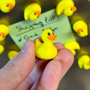 Puede incluir: Un peque&ntilde;o pato de goma amarillo brillante con pico naranja y ojos negros. El pato se sostiene en primer plano, mientras varios imanes de pato id&eacute;nticos est&aacute;n sujetos a una superficie met&aacute;lica, junto a una nota verde claro con el texto Shopping list.