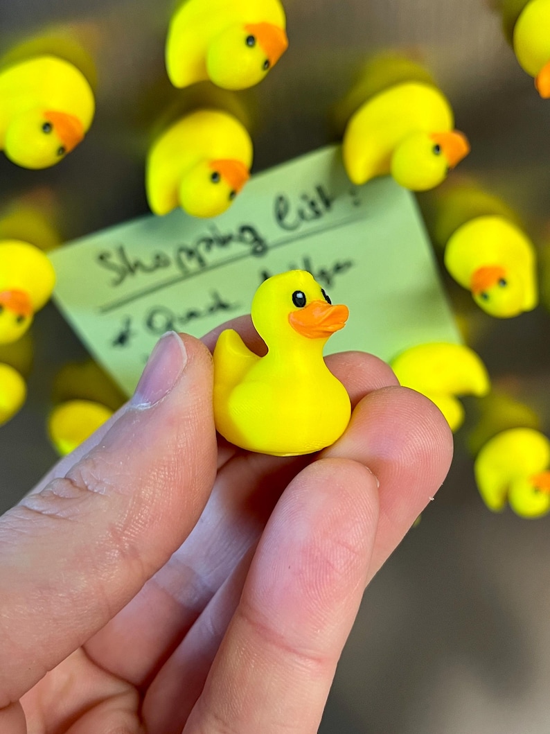 Puede incluir: Un peque&ntilde;o pato de goma amarillo brillante con pico naranja y ojos negros. El pato se sostiene en primer plano, mientras varios imanes de pato id&eacute;nticos est&aacute;n sujetos a una superficie met&aacute;lica, junto a una nota verde claro con el texto Shopping list.