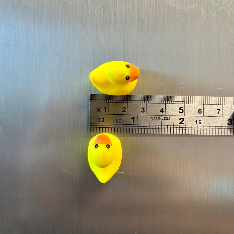 Puede incluir: Dos peque&ntilde;os patos de goma amarillos con picos naranjas y ojos negros. Una regla de acero inoxidable est&aacute; colocada entre ellos, mostrando que los patos miden aproximadamente 3,8 cm de largo desde el pico hasta la cola.