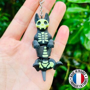 May include: A black and green skeleton dog keychain. The dog has a skull-shaped head and a rib cage design. The keychain is attached to a silver chain. The image also includes a "Made in France" sticker.