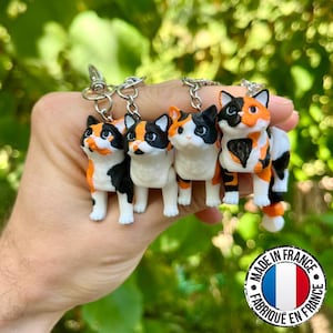 Peut inclure: Quatre porte-clés en forme de chat avec des marques orange, noires et blanches sont présentés. Chaque porte-clés a un fermoir en métal argenté. Les porte-clés sont alignés et les chats ont des motifs différents. Fabriqué en France.