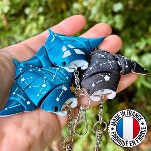 May include: Two articulated manta ray keychains, one in metallic blue and one in black, both featuring white constellation patterns on their dorsal sides. Each ray is attached to a silver-toned metal lobster clasp via a short chain link.