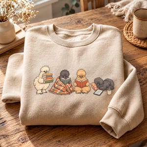 Può includere: Una felpa girocollo beige con un'illustrazione grafica di quattro soffici polli moroseta. Uno tiene in mano una pila di libri, un altro legge sotto una coperta a quadri, un terzo indossa occhiali mentre legge e il quarto dorme su un cuscino.
