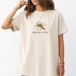 Può includere: T-shirt color crema con un disegno di zanzara dei cartoni animati. La zanzara ha grandi occhi e ali a strisce. Il testo "why am i so fly" è stampato sotto. La maglietta ha maniche corte e una vestibilità ampia.