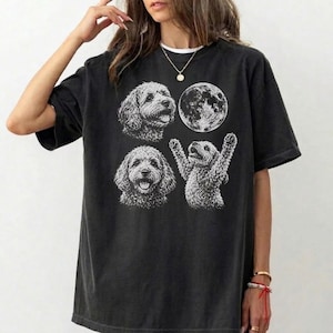 May include: A black short-sleeved t-shirt featuring a white graphic print. The design includes two illustrations of curly-haired dogs, one with its paws raised, and a detailed circular moon illustration positioned above the dogs on the chest area.