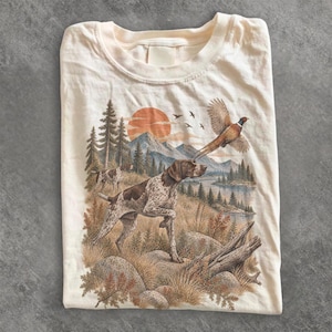 Könnte beinhalten: Ein cremefarbenes T-Shirt mit einem Grafikdruck eines braun-weiß gefleckten Jagdhundes, der auf einem grasbewachsenen Feld steht. Das Motiv zeigt zudem einen fliegenden Fasan, ein Gebirge, eine große orangefarbene Sonne und Nadelbäume.