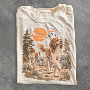 Puede incluir: Una camiseta de color crema con un estampado gráfico de dos perros tipo spaniel marrones y blancos en un paisaje. La escena incluye un gran sol naranja, nubes, pinos, un lago y una valla de madera. Los perros aparecen caminando en un campo.