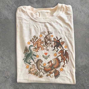 May include: A cream-colored t-shirt featuring a circular graphic print of various mythical creatures and animals, including a sasquatch, a mothman, a dragon, and a dinosaur, all dancing around a central red heart and small yellow stars.