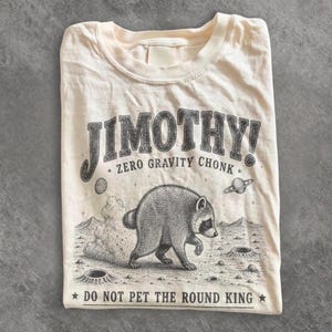 Könnte beinhalten: Ein cremefarbenes T-Shirt mit einer schwarzen Grafik eines Waschbären auf einer Mondoberfläche. Der Text lautet JIMOTHY! ZERO GRAVITY CHONK und DO NOT PET THE ROUND KING. Das Design enthält kleine Sterne, Planeten und Krater auf dem Mondboden.