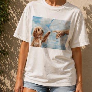 Könnte beinhalten: Ein weißes Kurzarm-T-Shirt mit einem rechteckigen Grafikdruck auf der Brust. Das Bild zeigt einen goldbraunen Welpen, der seine Pfote vor einem blauen Himmel in Richtung eines menschlichen Fingers ausstreckt, in Anlehnung an die Erschaffung Adams.