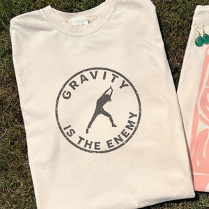 Puede incluir: Camiseta color crema con un gráfico circular que dice "GRAVITY IS THE ENEMY" en una fuente desgastada. El gráfico presenta la silueta de una persona en una postura de yoga. La camiseta está hecha de un material suave.