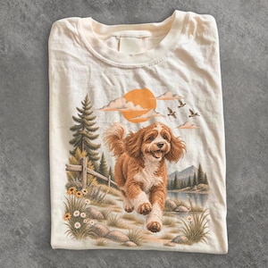 Peut inclure: Un t-shirt couleur crème avec une impression graphique représentant un chien marron courant dans un paysage composé de pins, d'une clôture en bois, de fleurs sauvages, d'un lac, de montagnes et d'un soleil orange avec des nuages et des oiseaux.