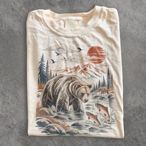 Könnte beinhalten: Ein cremefarbenes T-Shirt mit einer grafischen Illustration eines großen braunen Bären, der in einem Fluss watet. Die Szene zeigt zwei springende Fische, einen kleineren Bären in der Ferne, Tannen, Berggipfel und eine große orangefarbene Sonne.