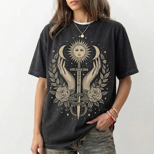 Può includere: T-shirt grigio scuro con un design celeste. La grafica include un sole, lune crescenti, mani, una spada, rose e stelle. La maglietta ha maniche corte e girocollo.