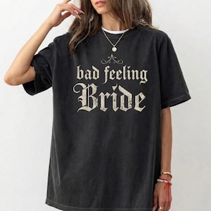 Puede incluir: Camiseta gris oscuro con el texto "bad feeling Bride" en una fuente vintage. La camiseta tiene cuello redondo y mangas cortas. La camiseta es de corte holgado.