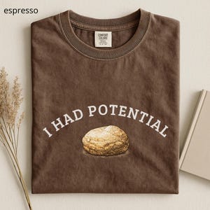 Può includere: T-shirt color espresso con la scritta bianca "I HAD POTENTIAL" ad arco sopra una grafica di biscotto. La maglietta ha un girocollo e un'etichetta Comfort Colors.