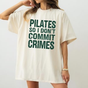 Può includere: T-shirt color crema con la scritta "PILATES SO I DON'T COMMIT CRIMES" in lettere verdi scuro. La maglietta ha maniche corte e una vestibilità rilassata, adatta per il tempo libero o lo sport.