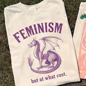 Può includere: T-shirt color crema con la scritta "FEMINISM" in viola arcuata nella parte superiore. Sotto, un'illustrazione di un drago viola. Il testo "but at what cost." è stampato sotto il drago.