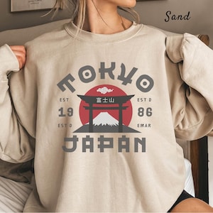 Puede incluir: Sudadera de color arena con un diseño gráfico que presenta la palabra "TOKYO" en gris, un círculo rojo, una ilustración de montaña y la palabra "JAPAN" en gris. La sudadera también tiene el texto "EST 1986 EMAR".