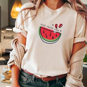 Può includere: T-shirt color crema con grafica a fetta di anguria e la scritta "MAKE PEACE LOUD". Il design dell'anguria include colori rosso, verde e nero. La maglietta è realizzata in un materiale morbido.