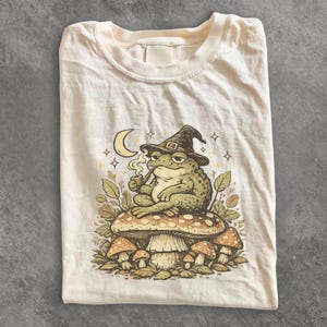 Può includere: Una t-shirt color crema chiaro con un'illustrazione grafica di una rana verde che indossa un cappello a punta, seduta su un grande fungo maculato mentre fuma la pipa. Sopra la rana sono raffigurati una luna crescente e piccole stelle.