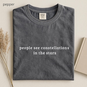 Può includere: Una maglietta grigia con la scritta bianca "people see constellations in the stars". La maglietta ha uno scollo tondo e un'etichetta con la scritta "Comfort Colors". La maglietta è piegata su una superficie beige chiaro.