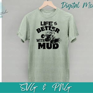 Op de afbeelding: Een groen T-shirt met de tekst "Life is better with mud" in zwart. Een zwart-witte illustratie van een persoon die een ATV door de modder rijdt, bevindt zich boven de tekst.
