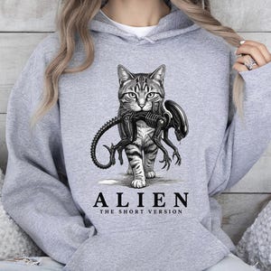 May include: A light gray hooded sweatshirt featuring a graphic print of a tabby cat carrying a small black alien creature in its mouth. Below the illustration, the text reads ALIEN THE SHORT VERSION in bold black capital letters.