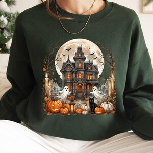 May include: A dark forest green long-sleeve sweatshirt featuring a central graphic print of a haunted mansion under a full moon, surrounded by flying bats, two white ghosts, a black cat, and several orange jack-o'-lanterns with lit candles.