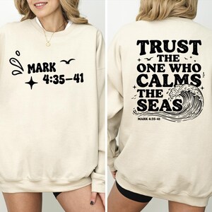 May include: A cream-colored crewneck sweatshirt featuring black graphic text. The front displays "MARK 4:35-41" with small bird and star icons. The back features the text "TRUST THE ONE WHO CALMS THE SEAS" above a large wave illustration.