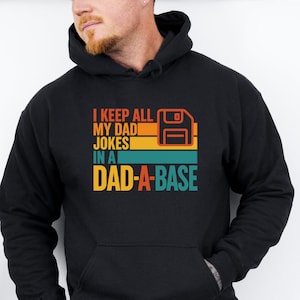 May include: Black hooded sweatshirt with the text "I KEEP ALL MY DAD JOKES IN A DAD-A-BASE" in orange, yellow, and teal. The design includes a retro floppy disk graphic. A casual, comfortable garment.