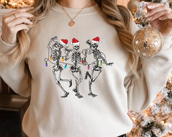 May include: Beige sweatshirt featuring a black and white illustration of three dancing skeletons. Each skeleton wears a red Santa hat and is strung with colorful Christmas lights. A festive and humorous holiday design.
