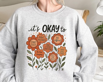 May include: A light gray sweatshirt with the text "it's OKAY to" above a floral design. The design includes orange flowers with phrases like "Start Over" and "Be Different".