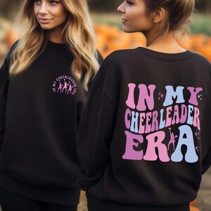 May include: Two black sweatshirts with a pink and blue graphic print. One sweatshirt says "In My Cheerleader Era" and the other says "In My Cheerleader Era".