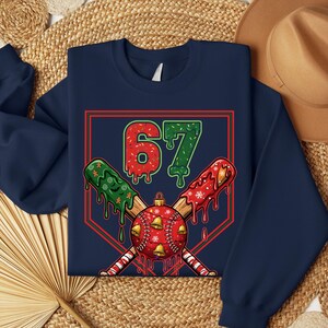 May include: Navy blue sweatshirt with a baseball-themed Christmas design. The graphic features the number 67 in red and green, crossed baseball bats, and a baseball with bells and snowflakes. The design is set within a red and green baseball diamond.