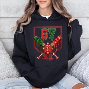 May include: Black hoodie featuring a baseball-themed Christmas design. The graphic includes crossed bats, a baseball ornament, and the number "67" in red and green. The design has a festive, holiday theme.
