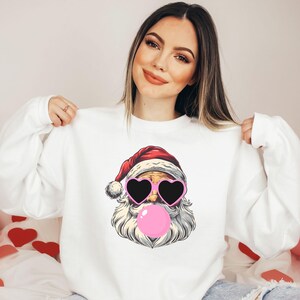 May include: White sweatshirt with a graphic of Santa Claus wearing pink heart-shaped sunglasses and blowing a pink bubble.